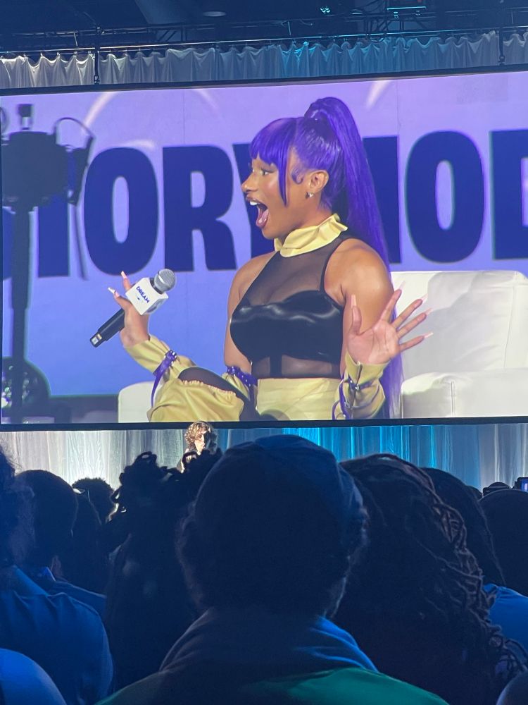 Megan Thee Stallion at DreamCon cosplaying as Yoruichi from Bleach. She is wearing a black mesh bodysuit, beige accessories around her neck, arms, and legs and has a purple high pony tail