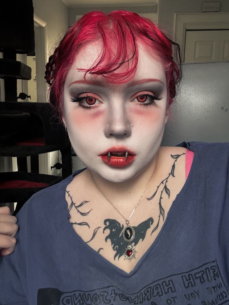 White foundation, sickly red tint to eyes and under eyes. Red irises. Pink and black hair in braids tied to head. Blue shirt with hat and vine tattoos visible on the chest 