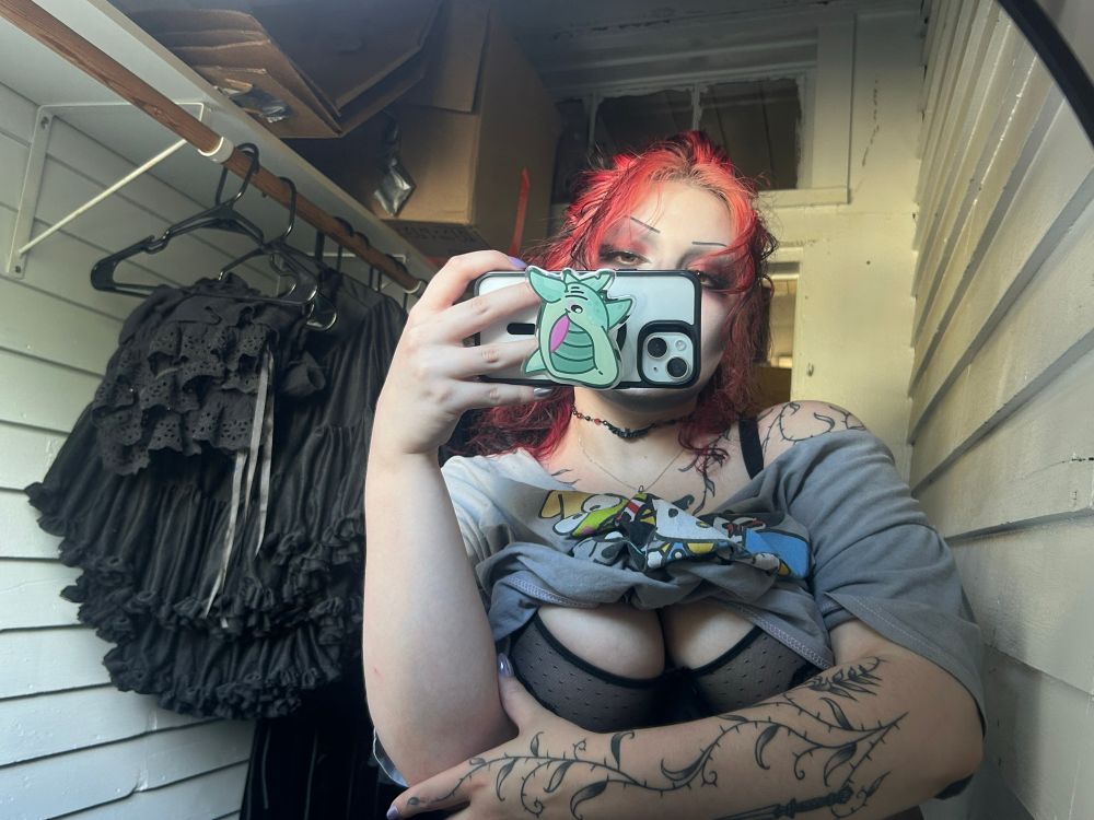Mirror selfie with black clothing in the background on hangers and shirt pulled up to expose bra