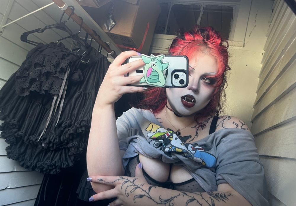 Mirror selfie with black clothing in the background on hangers and shirt pulled up to expose bra