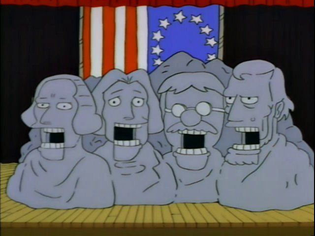 On a school assembly hall stage sits a papier-mâché Mount Rushmore prop with gaping ventriloquist dummy mouths for each head. The "Betsy Ross" flag hangs behind it.