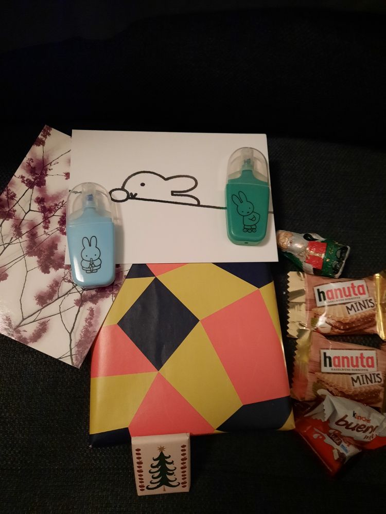 Ein in schwarz-gelb-orangenes Geschenkpapier verpackten Buch, eine Postkarte mit Weidenkätzchen als Aufdruck, eine zweite mit einem Häschen, das über eine Kante schaut; dazu zwei kleine Textmarker in grün und in blau, auf denen ebenfalls je ein kleiner Hase abgedruckt sind. Daneben liegen einige kleine Süßigkeiten.