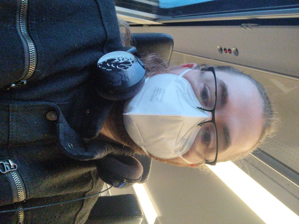 Ich sitze im Zug mit einer schwarzen Brille und FFP2 Maske im Gesicht.
An meinem Hals hängen Kopfhörer und ich trage einen schwarzen Mantel.
Links ist ein schmaler Streifen Fenster mit zartblauem Himmel zu sehen und über mir sind grelle Leuchtröhren