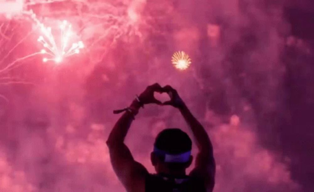 a man is making a heart shape with his hands while looking at fireworks .