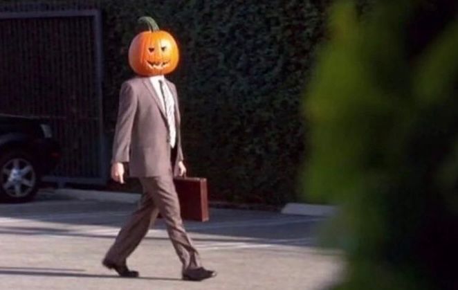 a still from the office: a man in a brown business suit is walking in a parking lot. he is holding a brown briefcase and wearing a jack o lantern on his head. 