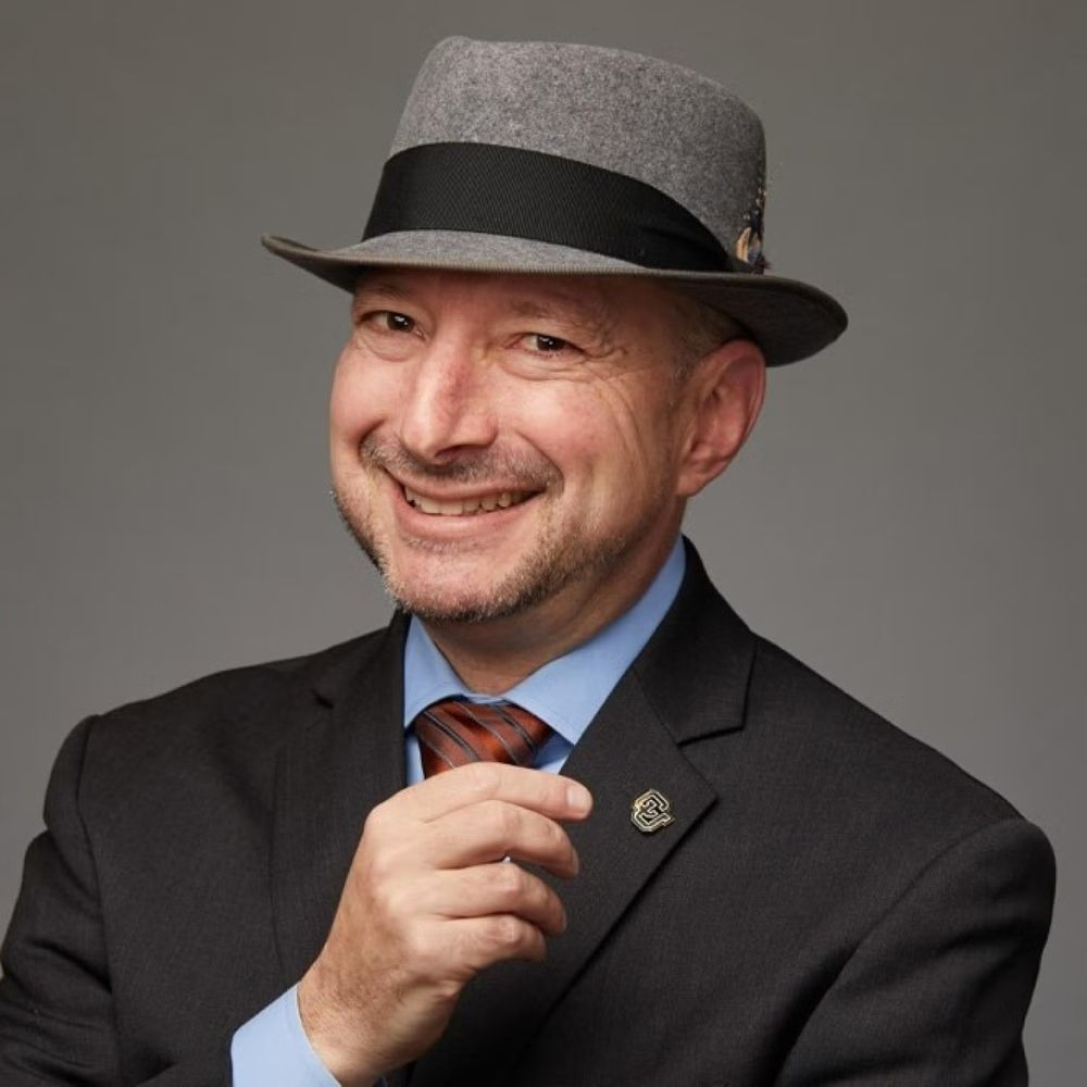 A headshot photograph of Dr. Finkelstein in a black sports coat with a UC lapel pin, red stripped tie, blue shirt, and grey fedora hat. 