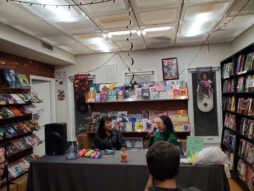 Two Korean women are sitting behind a table featuring the comic, The Fox Maidens. Behind them are shelves of indie comics.