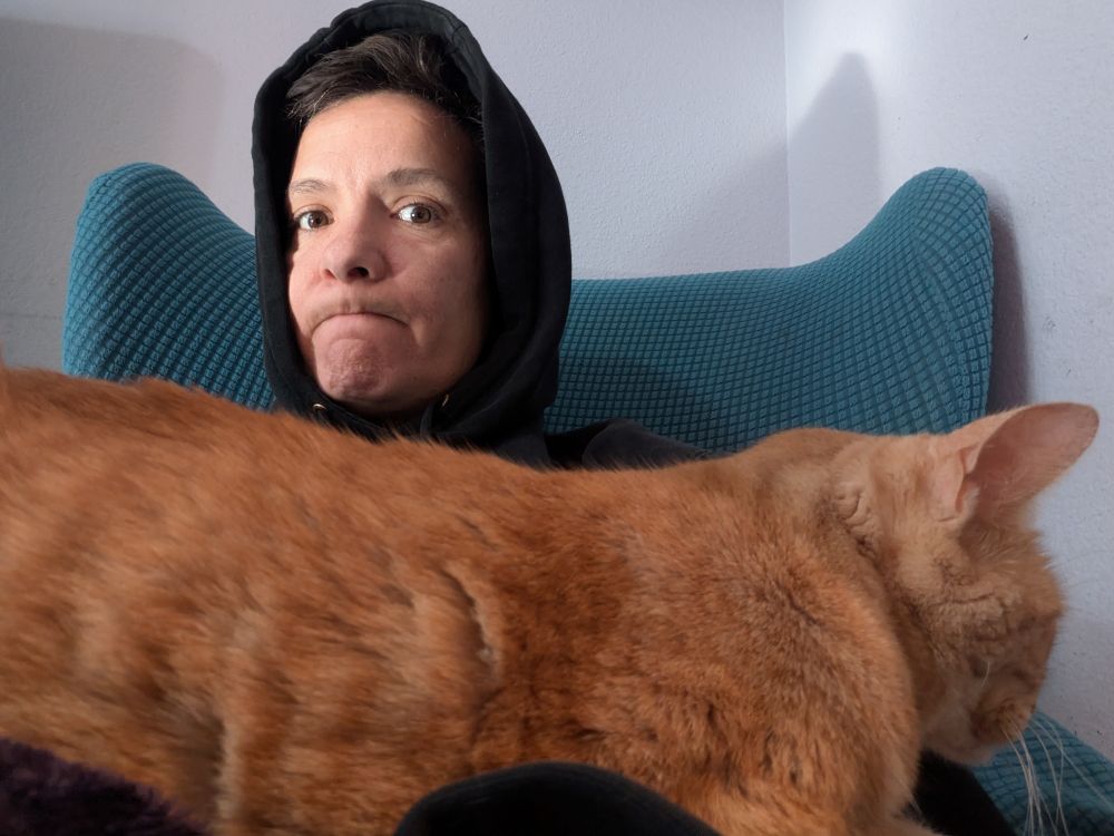 Selfie of me sitting in a blue armchair wearing a black hoodie with the hoodie up. Olive, an orange tabby cat, is stretched along my lap.