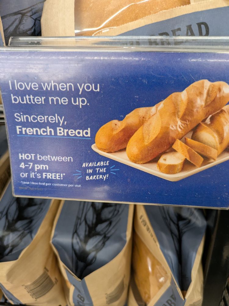 Photo of an advertising display inside a grocery store. The display is full of bags of bread. The sign says " i love when you butter me up. Sincerely, French Bread. HOT between 4-7 pm or it's FREE"