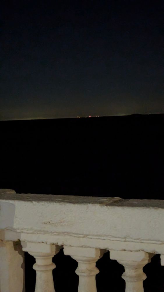 una foto vertical, más cerca a la cámara una baranda de cemento, y detrás el río de la plata, con luces brillando tenues a lo lejos 