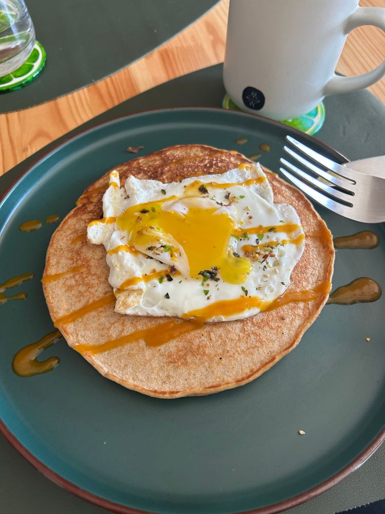 A savory pancake with a fried egg on top, the yolk has been broken. The pancake is on a green plate, with a green placemat in the background and a cup of coffee in the upper right hand corner.