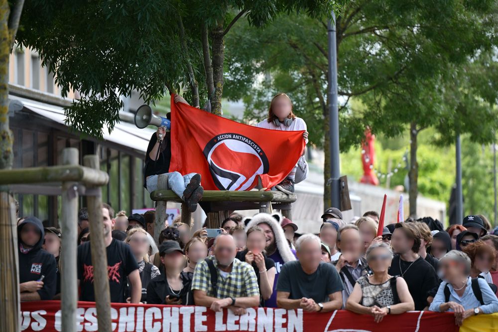 Auf einem Baum sitzen zwei Personen und halten eine Antifafahne. Darunter sind verschiedene Gegendemonstrierende zu sehen.