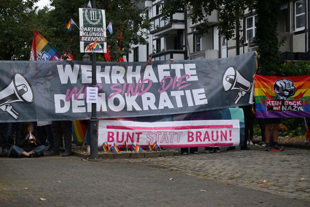 Es ist der Gegenprotest mit verschiedenen Transparenten zu sehen. Ein Wahlplakat des Der Dritte Weg wurde mit kleinen Prideflages verschönert. Auf einem großen Transparent steht "Wir sind die wehrhafte Demokratie". Auf einem weiteren "Bunt statt Braun".