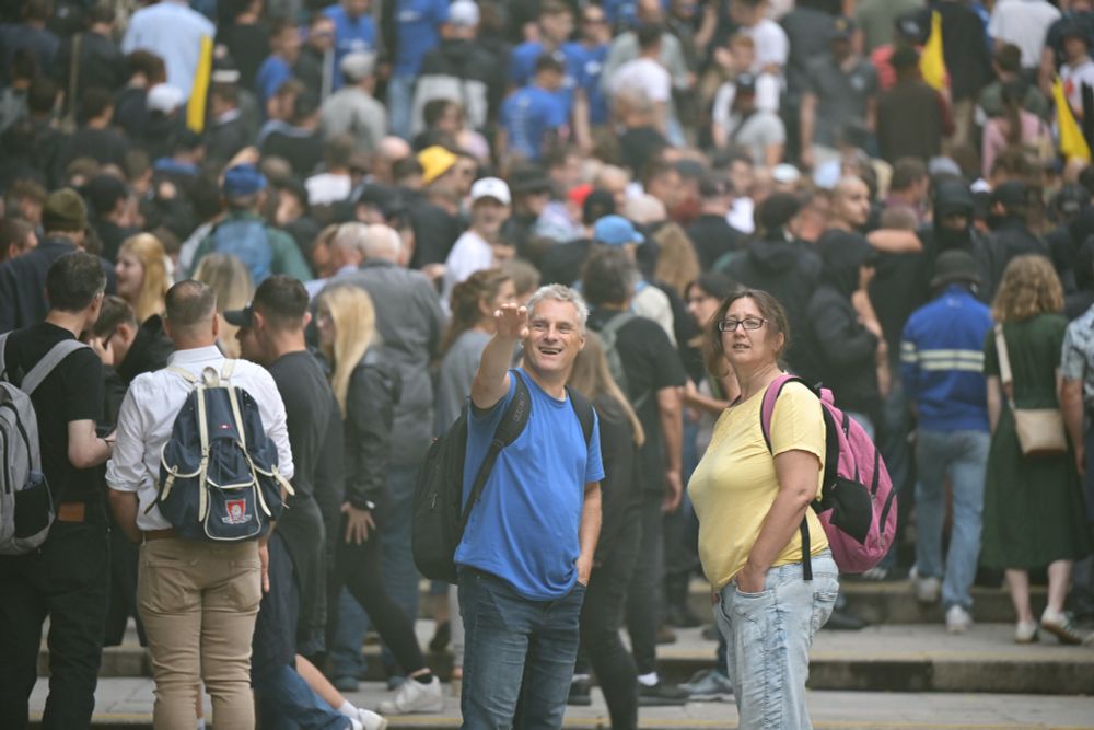 Ein Mann im blauen shirt hebt verdächtig seinen rechten Arm zum Hitlergruß. Daneben steht eine Frau. Im Hintergrund sind sehr viele Personen.