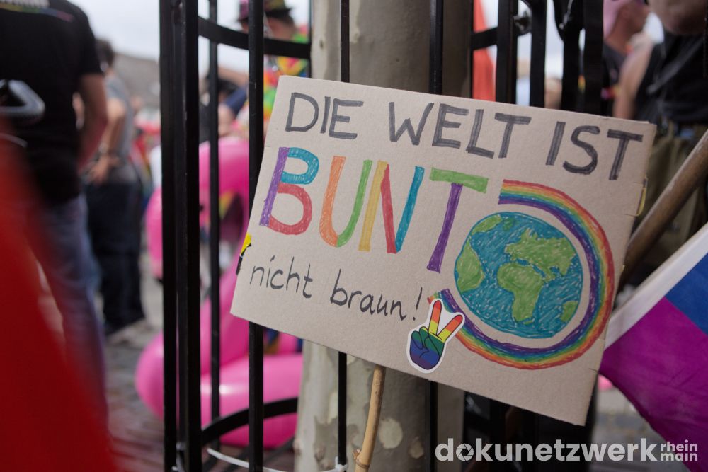 Ein Schild lehnt an einem Baum. Darauf steht: "Die Welt ist Bunt, nicht braun". Bunt ist in bunten Buchstaben geschrieben. Daneben ist eine Erde gezeichnet, die ein Regenbogen umgibt