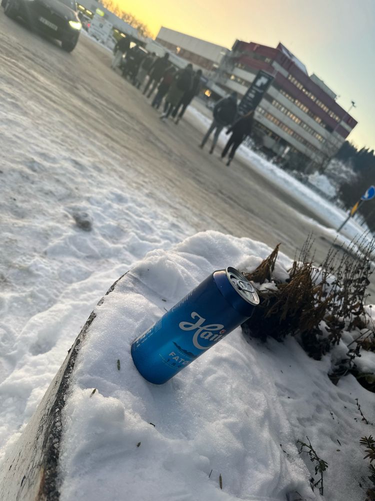 I front en forlatt Hansa fatøl i en snøfylt betongblomsterkasse,
I bakgrunnen skimtes NTNUs eksamenshus på Sluppen, en stor kloss av en bygning, og en lang rekke folk på vei mot bygget