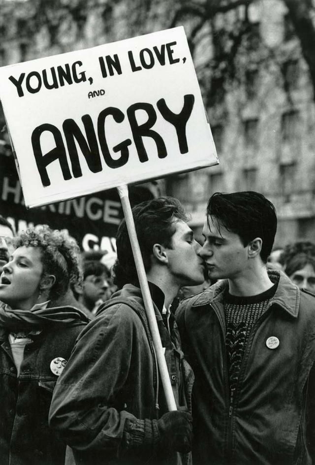 Black and white photo of a protest scene. In the foreground, two young men kiss while holding a sign that reads: “YOUNG, IN LOVE, AND ANGRY.” Both wear jackets and badges, and the mood is intimate and defiant. Behind them, other demonstrators are visible, one woman mid-chant. The image conveys themes of LGBTQ+ love, activism, and protest.