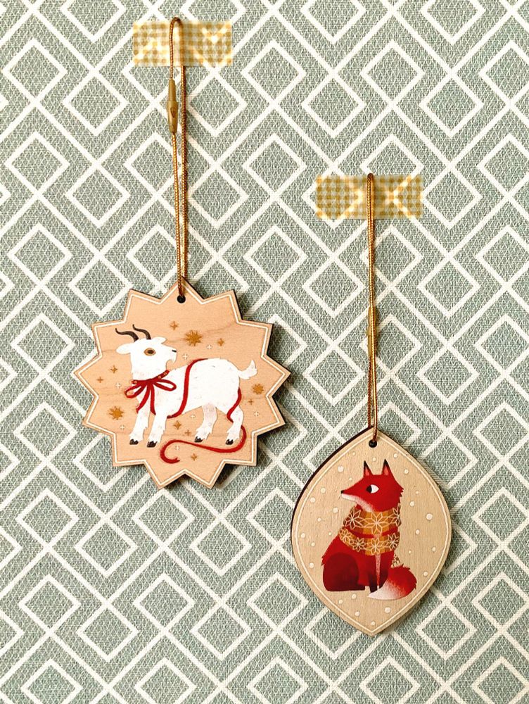 picture of two hanging wooden decorations: a star-shaped one with an illustration of a goat and a leaf-shaped one with a fox illustration
