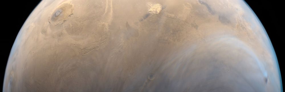 A high resolution photo of Mars from orbit captured by ESA's Mars Express. The image shows the reddish, dusty surface with soft brown and rusty hues, fading into a thin atmosphere. At the bottom left, two prominent volcanic formations, Olympus Mons and Ascraeus Mons, appear as large, broad features on the surface, with their edges softly blending into the landscape. On the bottom right, a large, circular bluish cloud hovers just above the surface, with soft, uneven edges. At the top center, a yellowish dust storm swirls across the terrain, resembling tightly packed cotton balls in a rough oval shape. The curvature of the planet highlights the thin atmosphere, gradually fading into the vast darkness of space.