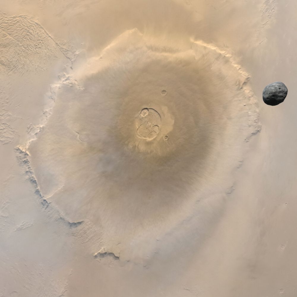 A high-resolution color image of Mars with the small, dark, potato shaped satellite Phobos, seen in sharp contrast as it passes over the massive volcano Olympus Mons. Phobos appears irregular and shadowed against the softly rust-colored Martian surface. Olympus Mons is a vast, circular shield volcano with gently sloping sides and a well-defined summit caldera at its center. Its broad base spans a large portion of the Martian surface, and subtle ridges can be seen radiating outward. The caldera appears slightly depressed, ringed by multiple nested collapse pits, giving it a rugged, textured look amid the surrounding smooth plains.
