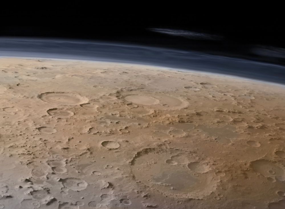 A digitally colourised image of Mars Promethei Terra region, captured by ESA’s Mars Express from orbit, shows a vast, cratered landscape near the massive Hellas Planitia basin. Dominating the scene are three major impact craters: Secchi Crater with a wide, sharply defined rim, Wallace Crater partially overlapping nearby craters and the more eroded Tikhov Crater with softened edges. The surface is textured with countless smaller craters, in tones of ochre, red, and dusty brown, evoking a parched desert. Above the horizon, a thin bluish layer of Mars atmosphere fades into deep black space. Wispy white clouds stretch across the upper layer of the atmospheric limb, offering a glimpse of Martian weather. The planet’s curvature is visible.