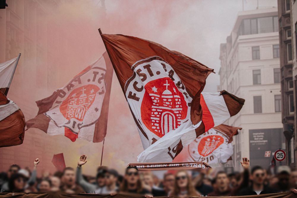 Eine Nahaufnahme von wehenden Fahnen des FC St. Pauli in den Vereinsfarben Braun, Weiß und Rot, mit dem Vereinslogo im Zentrum. Im Vordergrund jubeln Fans mit erhobenen Händen, während dichter Rauch und eine leicht verschwommene Stadtkulisse den Hintergrund füllen.