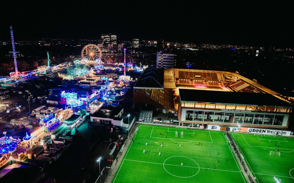 Aufnahme bei Nacht über St. Pauli: Links ein hell leuchtender Dom mit Riesenrad, bunten Fahrgeschäften und dicht gedrängten Wagen, rechts das ausgeleuchtete Millerntor-Stadion und die Trainingsplätze des FC St. Pauli, auf denen kleine Gruppen Fußball trainieren. In der Ferne die Stadtlichter.