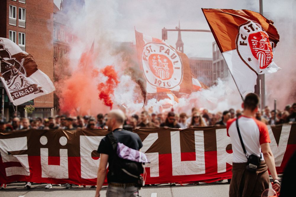 Eine Nahaufnahme von wehenden Fahnen des FC St. Pauli in den Vereinsfarben Braun, Weiß und Rot, mit dem Vereinslogo im Zentrum. Im Vordergrund jubeln Fans mit erhobenen Händen, während dichter Rauch und eine leicht verschwommene Stadtkulisse den Hintergrund füllen.