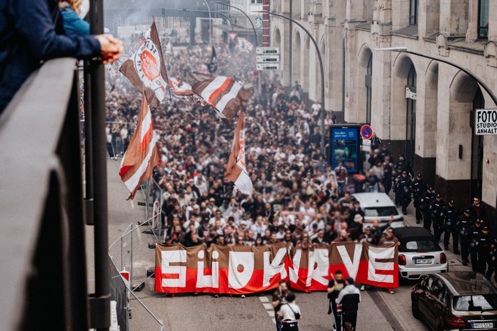 Ein Fanmarsch mit hunderten Menschen, die durch eine Hamburger Straße ziehen. Die vorderste Gruppe hält ein großes braun-rotes Banner mit dem Schriftzug „Südkurve“. Viele braun-weiße Fahnen mit dem Logo des FC St. Pauli wehen in der Luft. Auf beiden Seiten begleiten Polizisten den Zug.