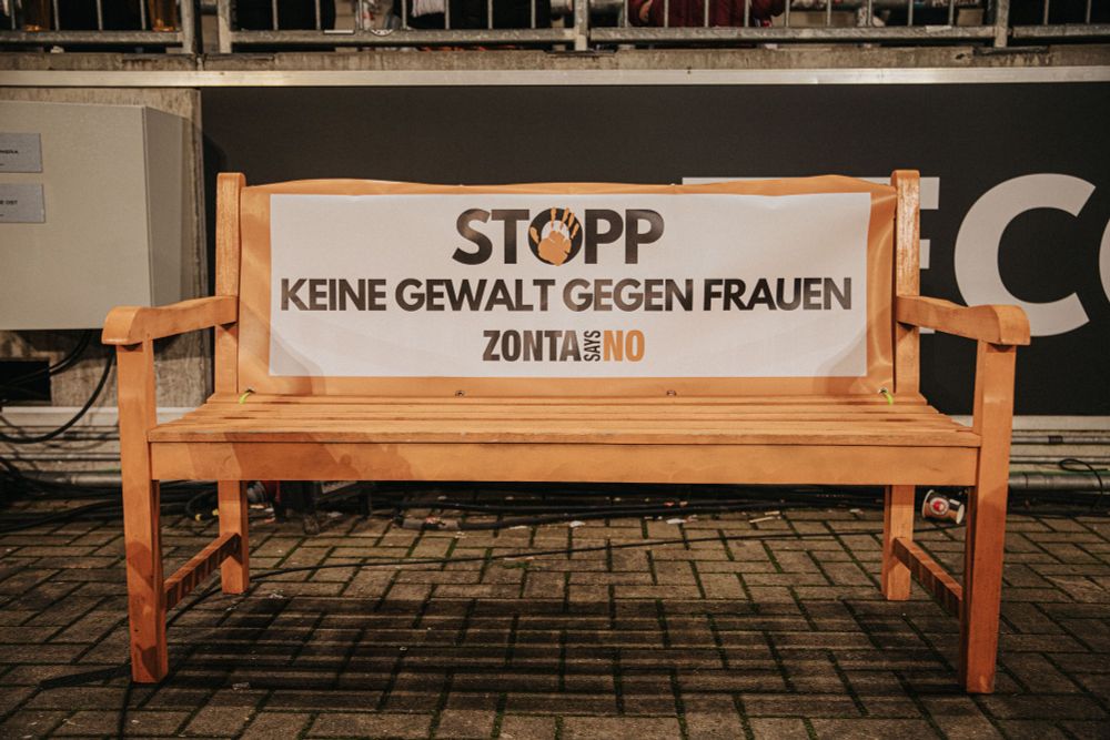 Das Bild zeigt eine orangefarbene Holzbank mit einem darauf befestigten Banner. Auf dem Banner steht in großen Buchstaben: "STOPP – KEINE GEWALT GEGEN FRAUEN". Darunter ist der Schriftzug "ZONTA SAYS NO" zu sehen. Es handelt sich um eine Aktion, die sich gegen Gewalt an Frauen richtet. Die Bank steht auf einem gepflasterten Untergrund vor einer Absperrung.