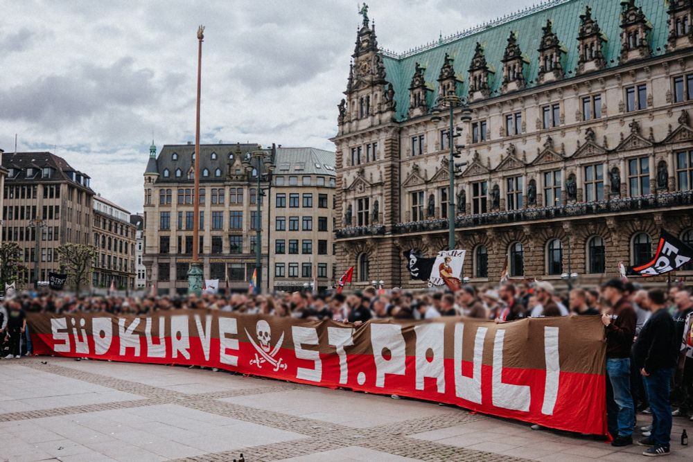 Eine große Menschenmenge steht vor dem Hamburger Rathaus und hält ein riesiges Banner mit der Aufschrift „Südkurve St. Pauli“. Auf dem Banner befindet sich auch ein Totenkopf mit gekreuzten Knochen – das Symbol des Fußballvereins FC St. Pauli. Im Hintergrund ist das imposante Gebäude des Rathauses mit grünem Dach zu sehen.