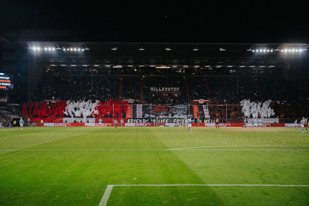 Das Bild zeigt die Südkurve im Millerntor beim Heimspiel gegen Union Berlin. Es wird eine Choreo gezeigt mit Fahnen auf der einen und braunen und weißen auf der anderen Seite. In der Mitte ein Spruchband mit den Logos beider Ultragruppen und der Aufschrift: Friends in Life - Friends in hell.