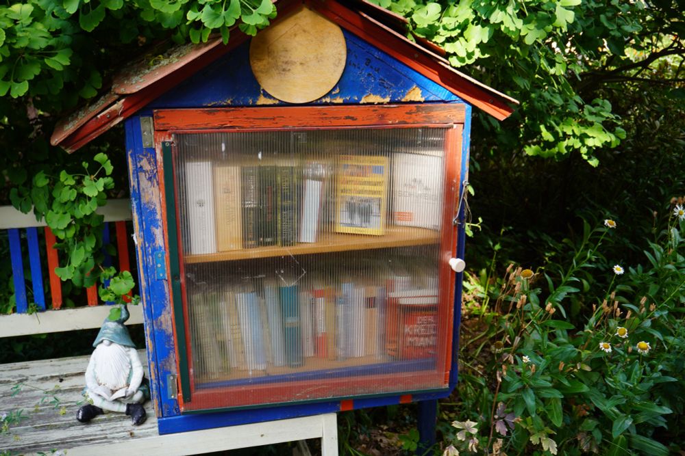 An very old little free library in blue and red with a lot of books and with a With the need to be renovated. And yes, there is a reading dwarf at it´s left side 