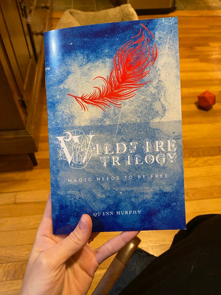 Somercon haul! A copy of the Wildfire Trilogy by Quinn Murphy. It’s a blue watercolory cover, dark on the top and bottom, light in the middle, with a red feather floating above the title, written in a pretty filigree font. Below the title says “magic needs to be free”