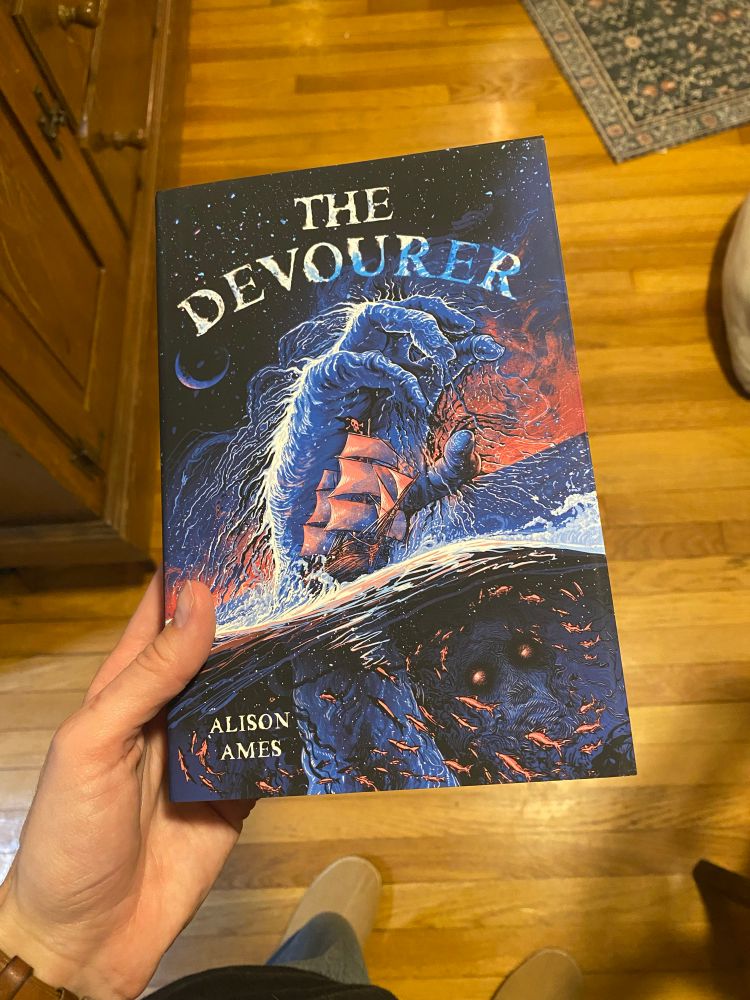 A book! The Devourer by Alison Ames. Blue and red colored cover art of a ship about to pulled into the ocean by a hand coming up out of the water, plus fish and creepy face under water