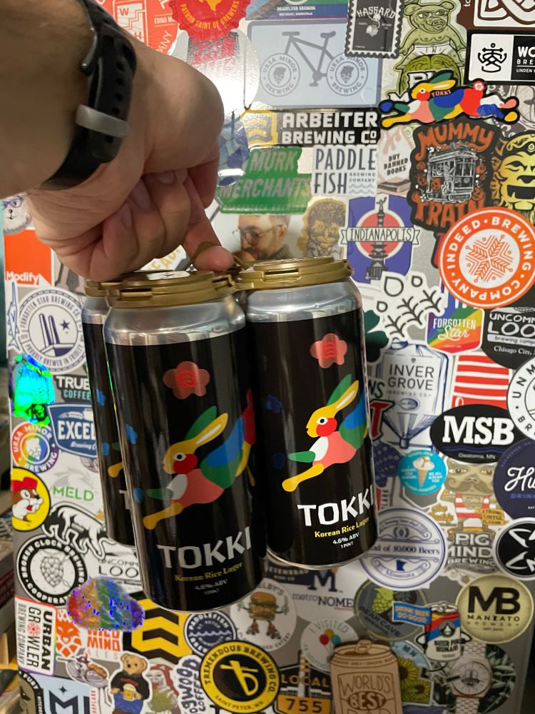 A 4-pack of TOKKI Korean rice lagers, raised up in front of a sticker-covered beer fridge