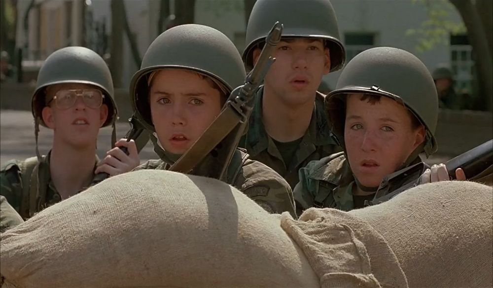 Kids with machine guns dressed up as soldiers behind sandbags. 