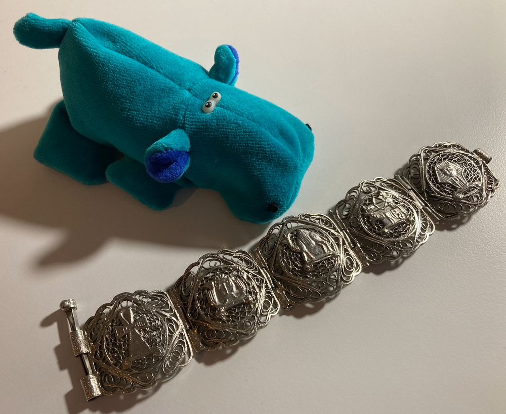 Little light blue hippo (stuffed toy) to the right and top, with darker blue ears. In front of Nils, there is a silver bracelet with five different links, showing an ankh, two uraei, a camel, pharaoh on a chariot, and (probably) the mask of Tutankhamun.