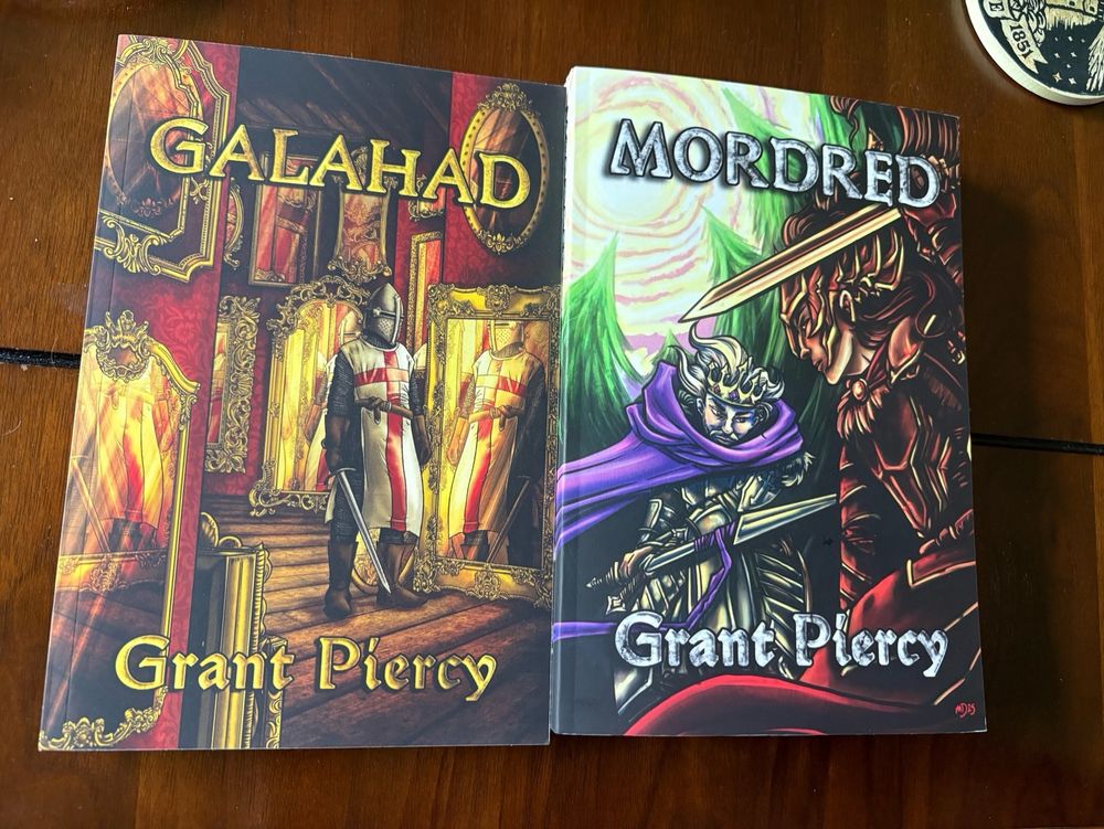 Physical copies of GALAHAD and MORDRED by Grant Piercy; cover art by Sanrixian. For GALAHAD a knight in red cross surcoat faces his own reflection in a palace of ornately decorated mirrors. For MORDRED, Sir Mordred in rust-colored armor charges at his father, King Arthur, wearing a crown of rubies and amethysts and a purple cloak.