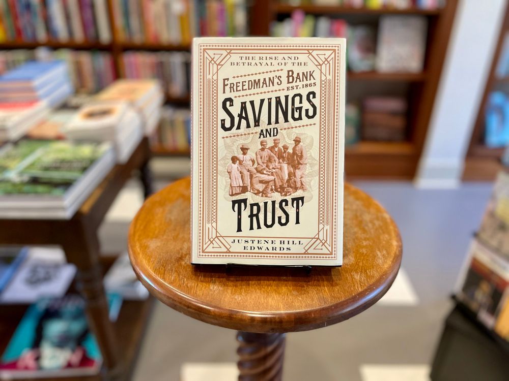A cream-colored book sits atop a small, round, wooden pedastal with a bookstore backdrop. The cover's frame mimics steel rust red in an art deco style of interlocking lines and patterns.

The cover reads: The Rise and Betrayal of the Freedman’s Bank Savings and Trust.

It's center is a photo of a group of Black men and children in various poses, mostly looking forward toward the reader.

The bottom of the cover names the author: Justene Hill Edwards. 