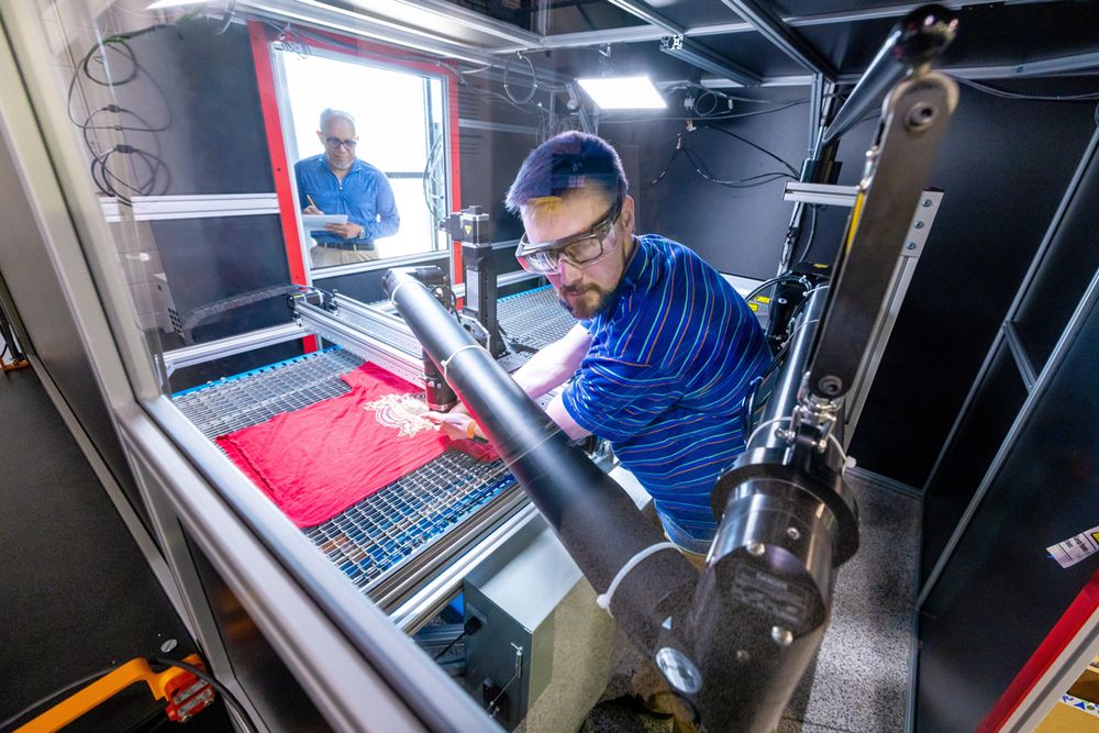 Two men are working on a machine. One is overseeing the work while another works on calibrating a robotic arm. I shirt is laying flat on a surface under metal arms and lasers