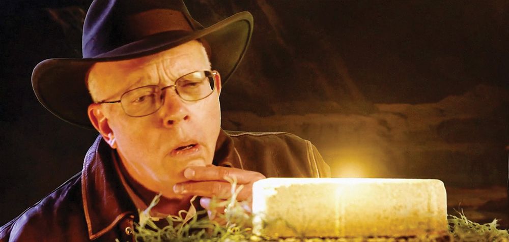 An older man with white hair is wearing glasses, a brown fedora, and a brown leather jacket while looking inquisitively at a shiny golden brick on a pedestal. It is meant to reference the Indiana Jones movies and character 
