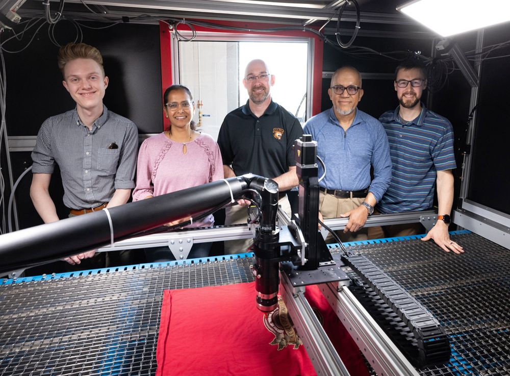 Group of five people pose next to a machine that has a flat table surface and a robotic arm with lasers