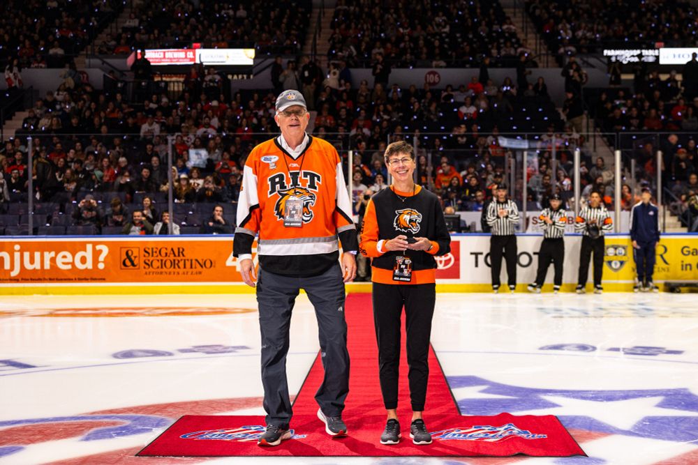 Older man and woman wearing R I T shirts walk down a red carpet on an ice rink in an arena holding a hockey puck