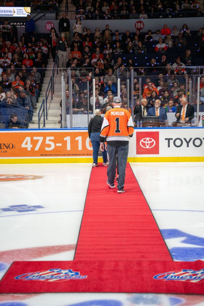 Older man wearing a baseball hat and hockey jersey walks down a red carpot off the ice in an arena at a hockey game


