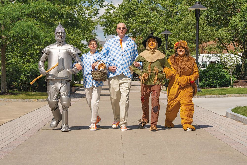 People dressed as characters from the Wizard of Oz movie are linked arm in arm while skipping down the Quarter Mile. Three students are dressed as the tin man, scarecrow, and lion. An older man and woman are dressed as a modern Dorothy in gingham blazers and orange sparkly converse sneakers 