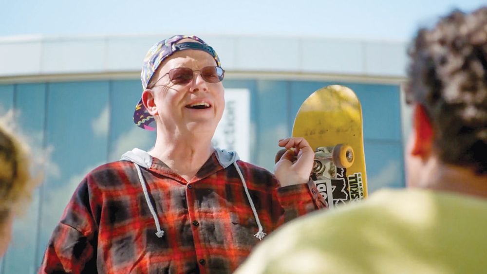 Older man with white hair is wearing a backwards baseball hat, glasses, and a hooded flannel shirt while holding a skateboard 