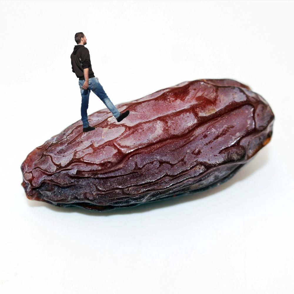 eine einzelne dattel (englisch: date) auf weißem hintergrund. auf ihr geht eine person mit jeans und schwarzem hoodie.