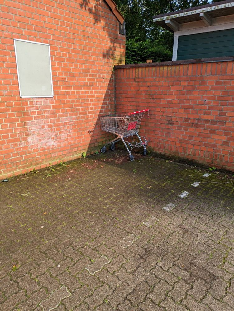Zwei Mauern im rechten Winkel auf einem Parkplatz von Edeka. In der Ecke steht ein Einkaufswagen von Rewe mit dem "Gesicht" zur Wand. Die Mauer legt einen Schatten über den Wagen. Sonst ist dort nichts weiter.