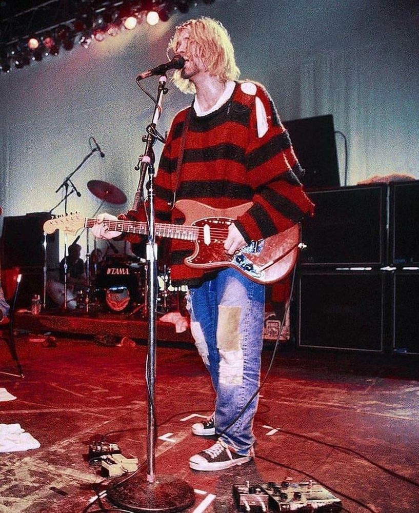 Kurt Cobain on a red stage wearing a black and red sweater and playing a red guitar. 